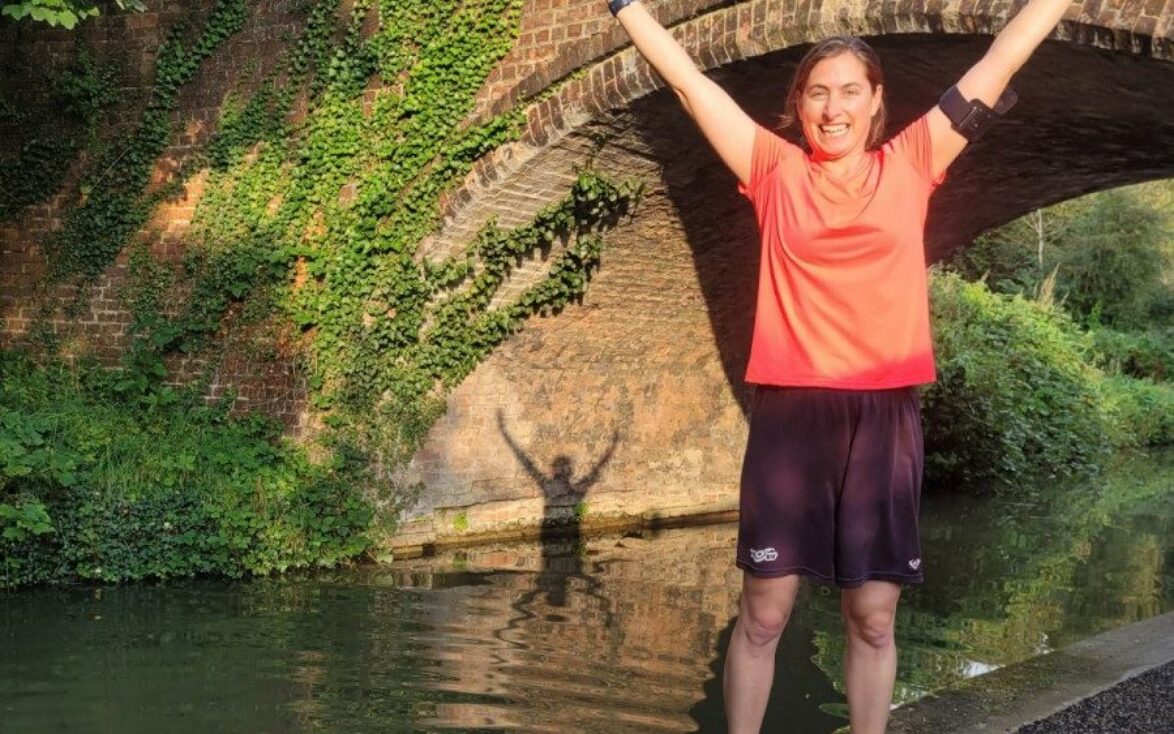 A woman in running gear stood by a canal, arms raised triumphantly.