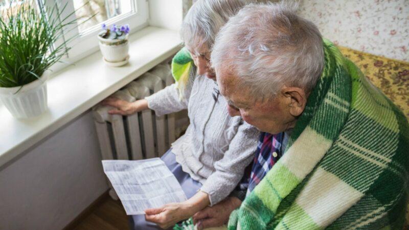 An elderly couple wrapped in a blanket feel their radiator while looking at a bill.