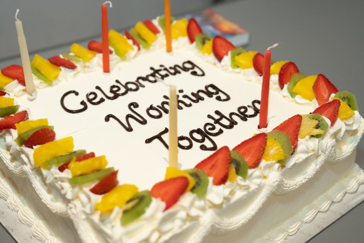 A cake with white frosting, decorated with colourful fruit with 'Celebrating working together' written in icing on the top