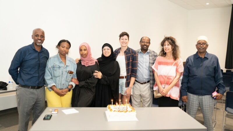 A group of people standing above a table with a cake and candles, smiling, to celebrate a successful partnership