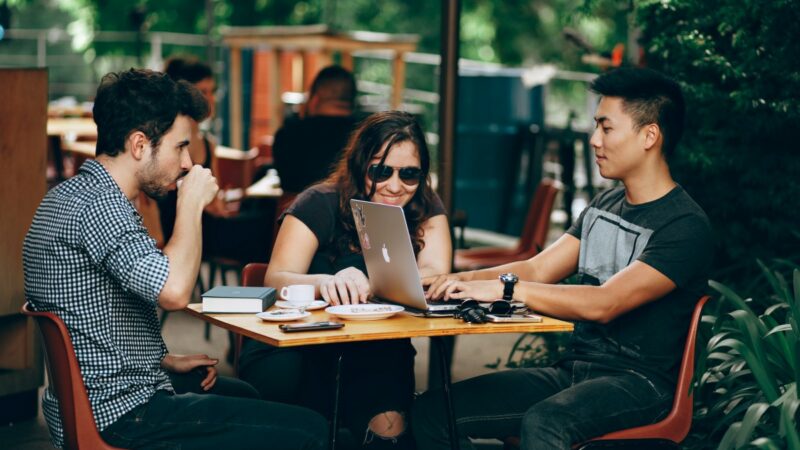 A group of three people sitting around a table in an outdoor cafe table. The are talking and working on a laptop.