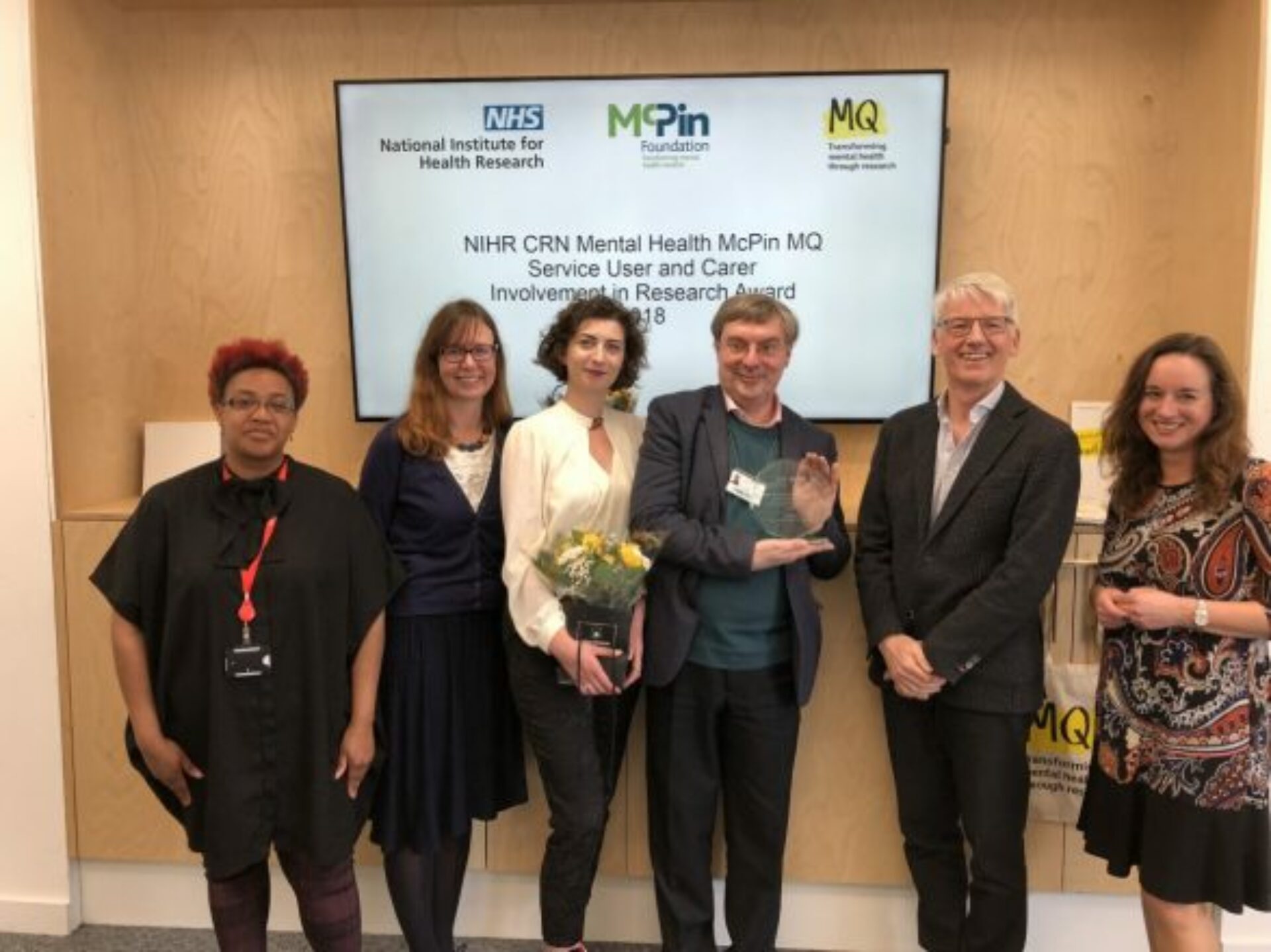 A group of people in front of a screen which reads 'NIHR CRN Mental Health McPin MQ Service User and Carer Involvement in Research Award 208'