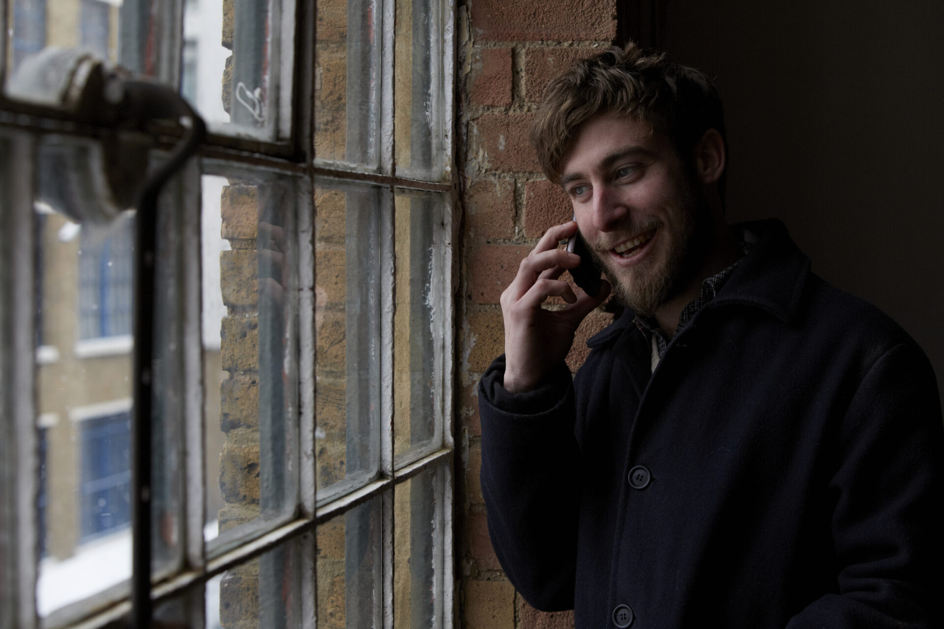 A man smiling and talking on a phone while looking out a window